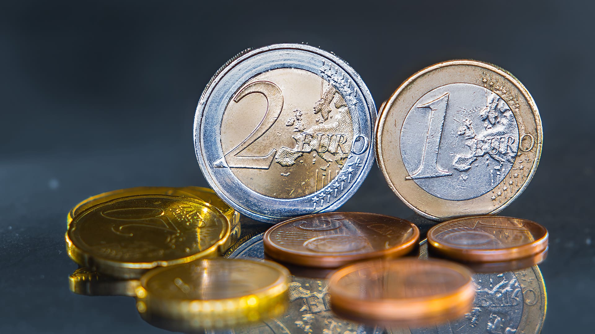 Vor den stehenden 1- und 2-Euro-Münzen liegen Cent-Stücke auf blau-grauen Hintergrund Vor den stehenden 1- und 2-Euro-Münzen liegen Cent-Stücke auf blau-grauen Hintergrund