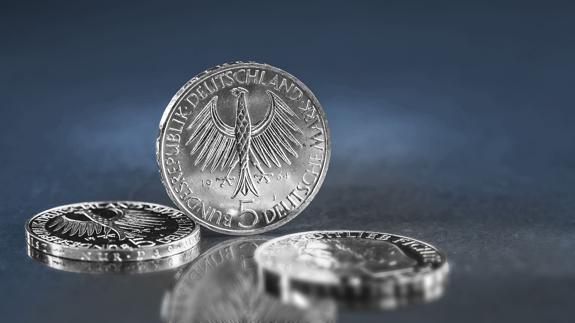 Eine 5-Mark-Münze steht zwischen zwei liegenden Mark-Münzen und zeigt dabei eine ihrer Seiten Eine 5-Mark-Münze steht zwischen zwei liegenden Mark-Münzen und zeigt dabei eine ihrer Seiten