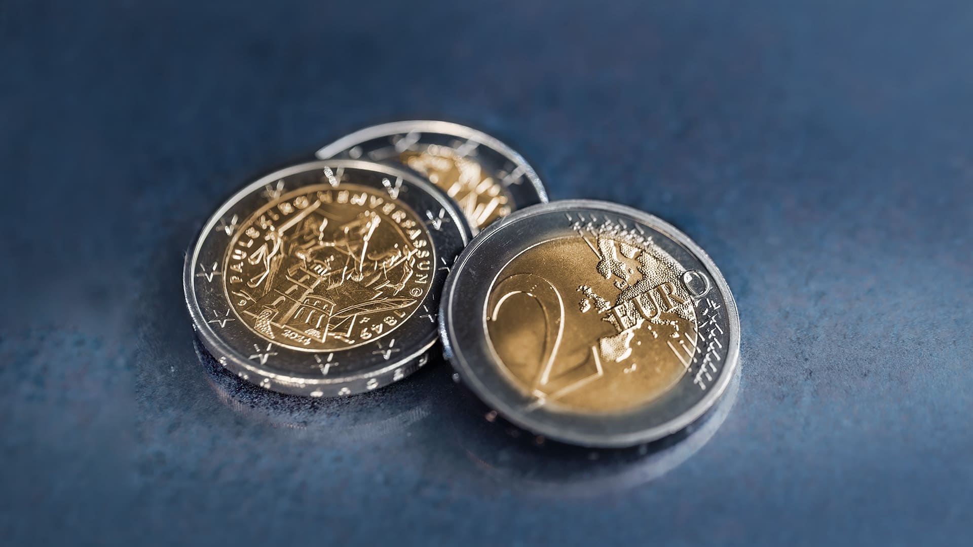 Drei 2-Euro-Münzen liegen auf einen blau-grauen Hintergrund Drei 2-Euro-Münzen liegen auf einen blau-grauen Hintergrund